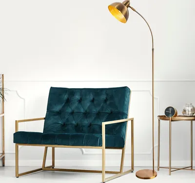 A teal velvet armchair with a gold frame is placed next to a gold floor lamp and a small side table with a clock and a glass vase.