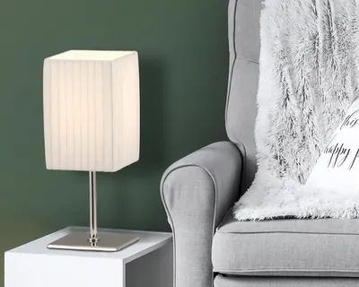 A table lamp sits on a white side table next to a gray armchair with a fluffy gray blanket draped over it.