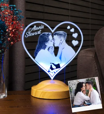 A heart-shaped personalized photo lamp with a couple kissing is displayed on a wooden base, with a vase of dried flowers and another framed photo in the foreground.