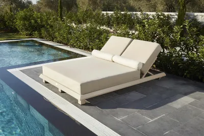 A beige sun lounger is placed on a stone patio next to a swimming pool.