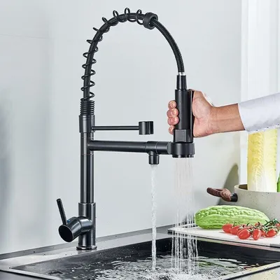 A black kitchen faucet with a pull-down sprayer is shown in use, with water running into a sink. Vegetables are visible on a cutting board in the background.