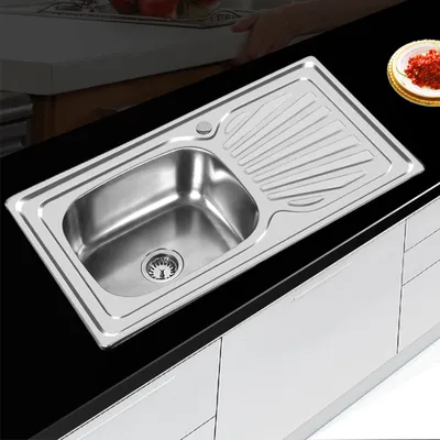 A stainless steel kitchen sink with a draining board is shown on a black countertop, with white cabinets below.