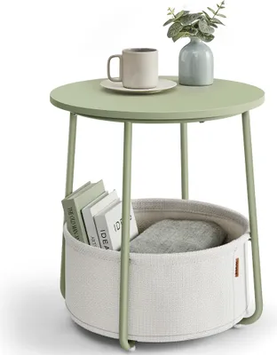 A round side table with a storage basket underneath, holding books and a grey cushion. A mug and a vase with greenery are on top.
