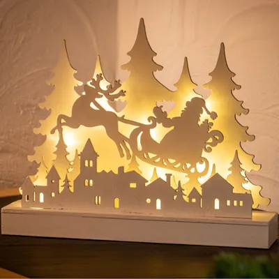 A lit-up wooden silhouette of Santa Claus in his sleigh being pulled by reindeer over a village, with Christmas trees in the background.