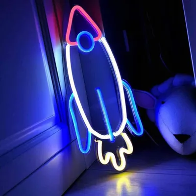 A neon rocket sign is displayed in a room, casting colorful light on the wall and floor. A stuffed animal is visible in the background.