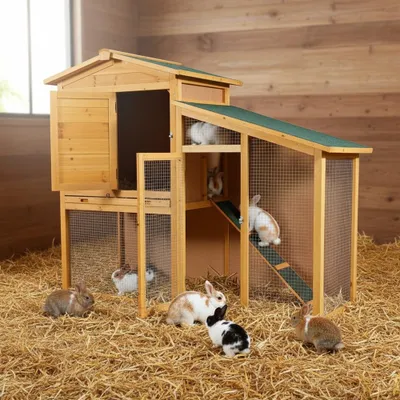 A wooden rabbit hutch with multiple levels and doors, filled with straw, and surrounded by several rabbits.