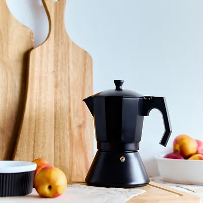 A black moka pot is displayed on a wooden surface with cutting boards and fruit in the background.