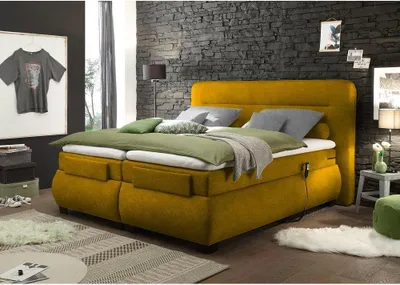 A bedroom scene featuring a yellow boxspring bed with green and gray bedding, set against a dark stone wall.