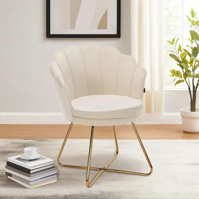 A beige velvet accent chair with gold metal legs is placed on a rug in a room with a framed picture on the wall and a plant in the background.