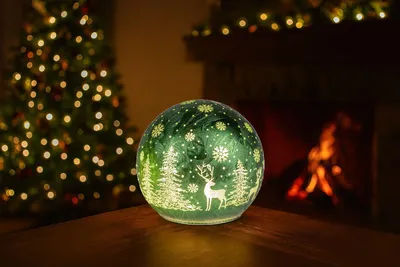 A decorative LED glass ball with a Christmas theme sits on a wooden surface in front of a fireplace and a Christmas tree.