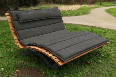 A chaise lounge with a gray cushion is placed on grass next to a winding path in a park.
