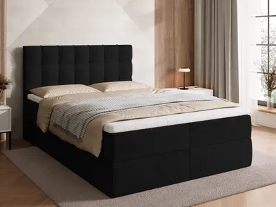 A black upholstered box spring bed with a tufted headboard, made up with white and tan bedding, is shown in a bedroom setting.