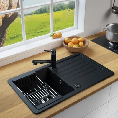 A kitchen sink with a faucet, a dish rack, and a bowl of oranges on a wooden countertop, with a window overlooking a green landscape in the background.