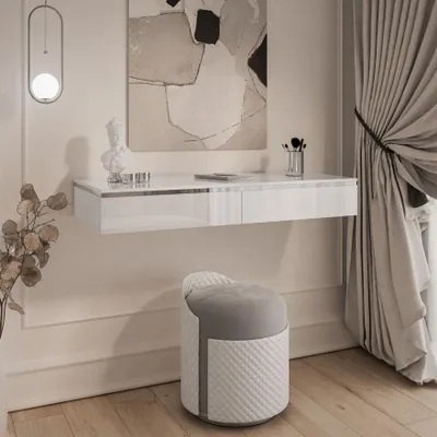 A modern white high-gloss floating vanity table with a decorative bust and makeup brushes on top, paired with a quilted stool in front of a wall with abstract art and curtains.