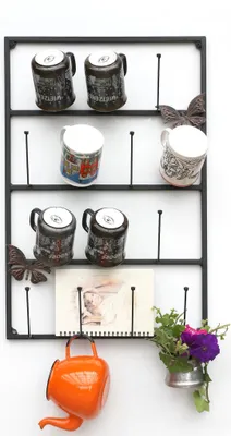 A black metal mug rack with several mugs, decorative butterflies, a small notepad, an orange teapot, and a small vase with flowers.