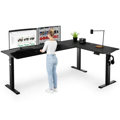 A woman stands at a black L-shaped standing desk with two monitors, a lamp, and headphones.