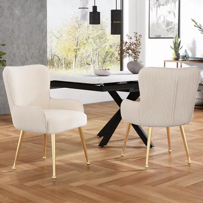 Two beige dining chairs with gold legs are placed on a herringbone wooden floor in front of a dining table. A large window with a view of trees is in the background.