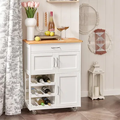 A white kitchen cart with a wooden top, stocked with wine bottles, sits next to a window with curtains. Decorative items like a vase of tulips, a candelabra, and wall art are also visible.