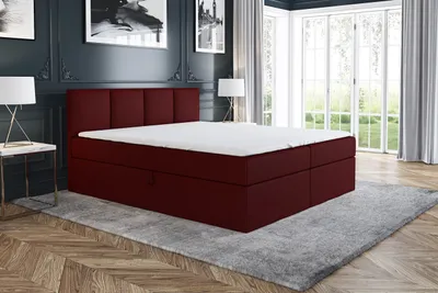 A red boxspring bed with a white mattress and topper is displayed in a bedroom setting with dark blue walls, a gray rug, and large windows in the background.