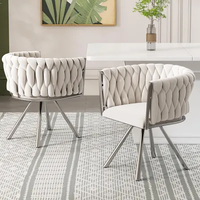 Two beige upholstered dining chairs with silver legs are placed on a grey and white striped rug in front of a white dining table.