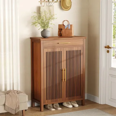 A wooden shoe cabinet with slatted doors and a drawer, placed in an entryway next to a door and a bench with a blanket.