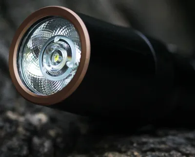 Close-up of the head of a black flashlight with a copper ring around the lens, resting on a textured surface.