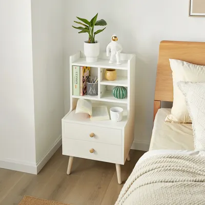A white nightstand with drawers and shelves is placed next to a bed.