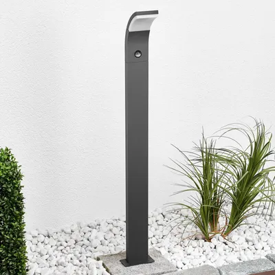 A modern, dark gray LED pathway light stands in a garden setting with white gravel and green plants.