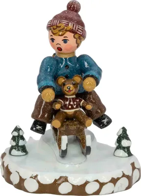 Ein fröhliches Kind mit blauer Jacke und roter Mütze sitzt auf einem Schlitten und hält einen Teddybären während es von kleinen Tannenbäumen umgeben auf einer schneebedeckten Basis platziert ist