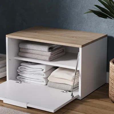 A white cabinet with a wood-effect top, open to reveal neatly folded white and gray textiles on two shelves. The cabinet is placed on a wooden floor against a dark blue wall.