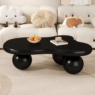 A black coffee table with a unique, organic shape sits on a cream-colored rug in front of a white sofa. The table has a bowl of fruit and a book with a cup on it. Decorative pillows, including a cloud-shaped one and a round one, are on the sofa.