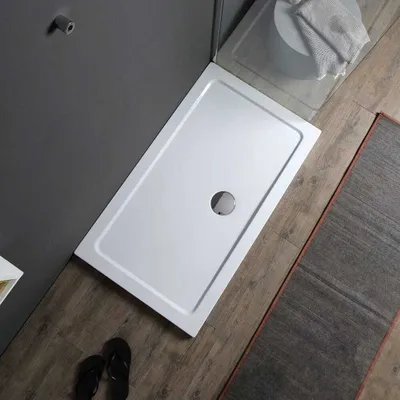 A rectangular acrylic shower tray is shown in a bathroom setting, with a gray wall, wooden floor, and a rug.