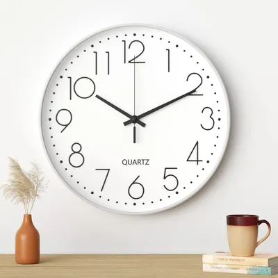 A white analog wall clock with black numbers and hands is mounted on a white wall. A small brown vase with dried grass is to the left of the clock, and a stack of books with a mug on top is to the right.