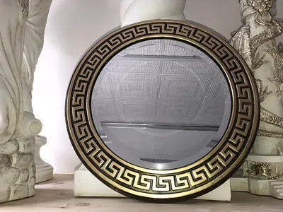 A round mirror with a Greek key pattern frame is displayed on a shelf, flanked by two decorative statues.
