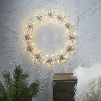 A circular LED light decoration resembling a flower ring is mounted on a textured white wall. In the foreground, a pine branch and a rustic wooden crate are visible, along with a fluffy white cushion.