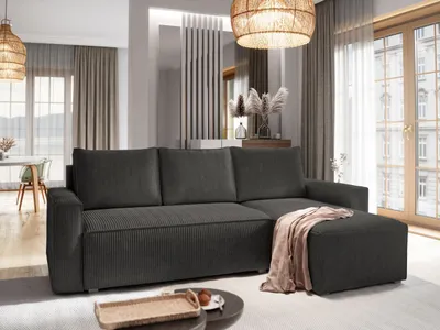 A modern living room with a dark gray sectional sofa and an ottoman, decorated with a beige throw blanket. The room features natural light from large windows and a woven pendant light fixture.