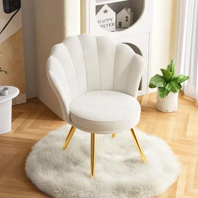 A white velvet accent chair with gold legs sits on a fluffy white rug in a room with wooden floors and a white bookshelf in the background.