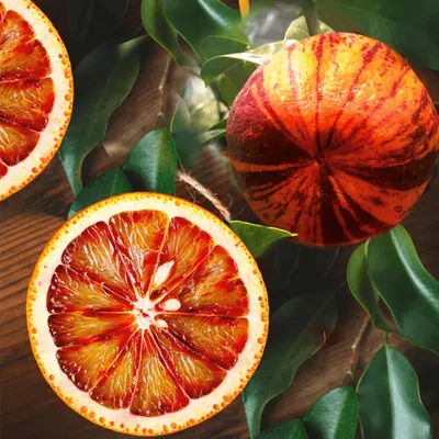 A close-up view of a sliced blood orange and a whole blood orange, surrounded by green leaves, on a wooden surface.