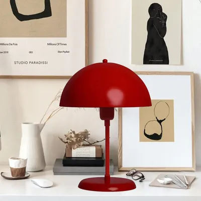 A red mushroom-shaped desk lamp is placed on a white desk next to a white vase, books, and framed artwork.