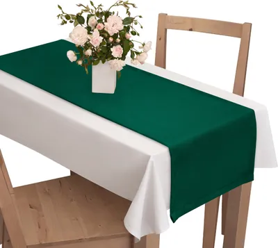 A dark green table runner is laid on a white tablecloth on a wooden table, with a vase of pink flowers on top.