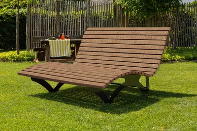 A wooden chaise lounge is placed on a green lawn in a garden setting. In the background, there is a wooden table with drinks and a striped cloth.