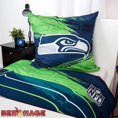 A bedding set with a Seattle Seahawks theme is displayed on a bed, with a nightstand and lamp in the background.