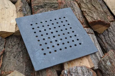 A square cast iron grate with many holes is placed on a pile of firewood.