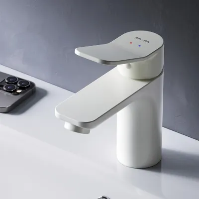 A white faucet is shown on a white sink, with a gray wall in the background. A smartphone is also visible on the left side of the image.