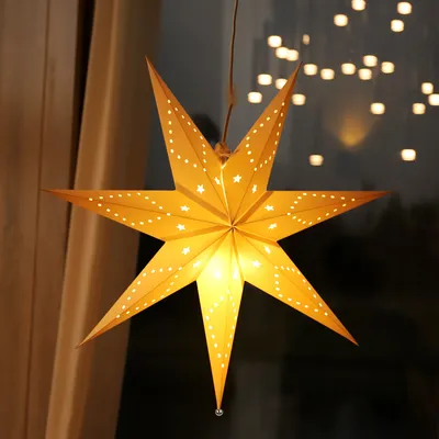 A yellow paper star lantern with small white star and dot cutouts is hanging in front of a window with blurred lights in the background and curtains to the side.