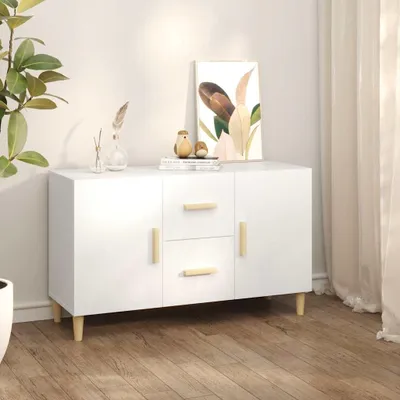A white sideboard with wooden legs and handles, decorated with a painting and small decorative items, placed on a wooden floor next to a plant and curtains.