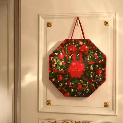 An advent calendar shaped like a wreath, decorated with Christmas ornaments and bows, hanging on a white door.
