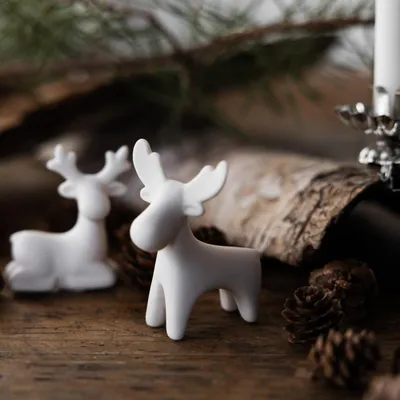 Two white ceramic reindeer figurines are placed on a wooden surface with pinecones and greenery in the background.