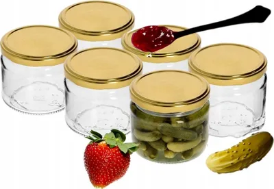 A group of empty glass jars with gold lids, one filled with pickles, a strawberry, and a pickle on a white background.