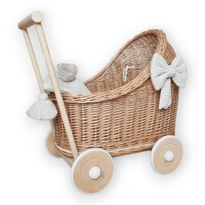 A wicker doll's pram with wooden handles and wheels, decorated with a white bow and tassels, and containing a grey knitted blanket and toy.
